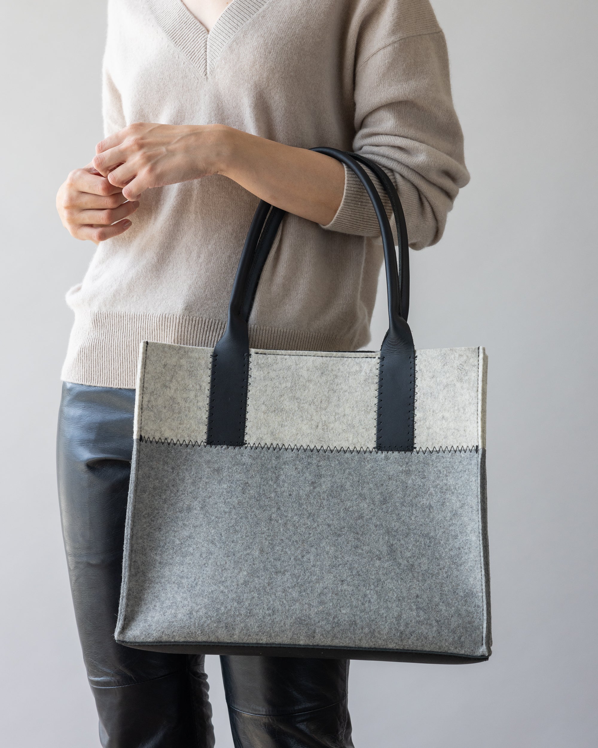 With a neutral backdrop, someone carries the chic Jaunt Midi Tote by graf lantz, a stylish two-toned gray felt bag with black leather handles while wearing a beige sweater and black leather pants.