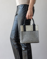A person in black leather pants holds a stylish graf lantz handbag made from luxurious Merino wool featuring black handles, beautifully contrasted against a neutral gray background.