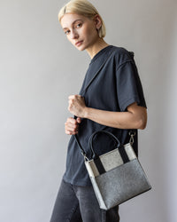 A person with short blond hair stands against a plain background, holding a versatile graf lantz handbag in gray and black. They wear a loose-fitting dark gray T-shirt and black jeans, looking toward the camera with a neutral expression.