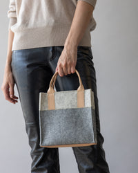 The person holds a versatile graf lantz Jaunt Mini Merino Wool Tote, made from sustainable materials in two-toned gray and beige felt with leather handles. They pair it effortlessly with a beige sweater and black leather pants, standing against a plain background.