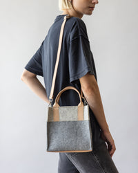 A person with short blonde hair, dressed in a dark t-shirt and jeans, carries the stylish and versatile graf lantz Jaunt Mini Merino Wool Tote on their shoulder. The tote, crafted from sustainable materials with distinctive beige handles, stands out against a plain background.