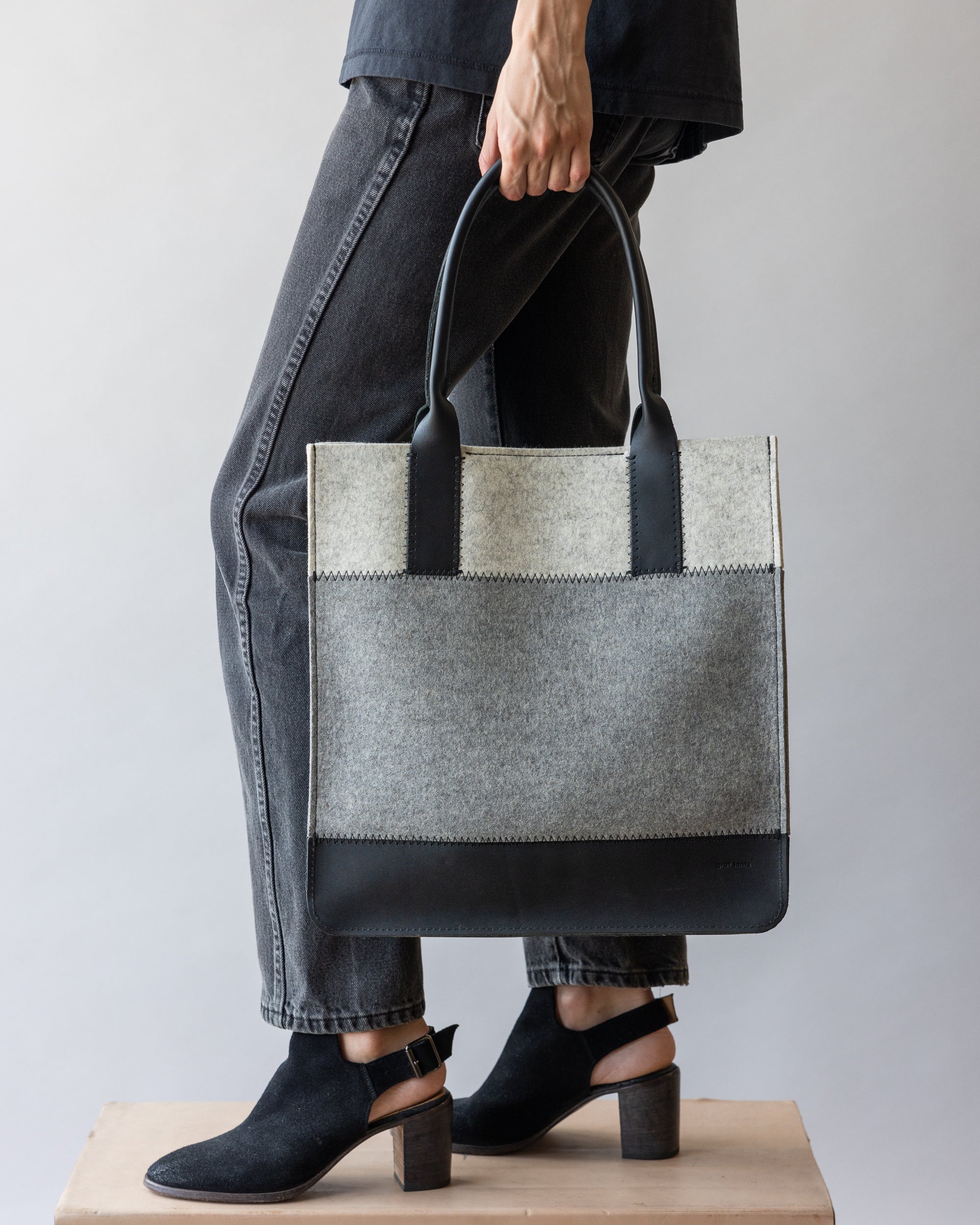 A person in dark jeans and black shoes holds a large, stylish graf lantz Merino Wool Tote with a gray fabric body and black leather handles. The setting is neutral and minimalistic.