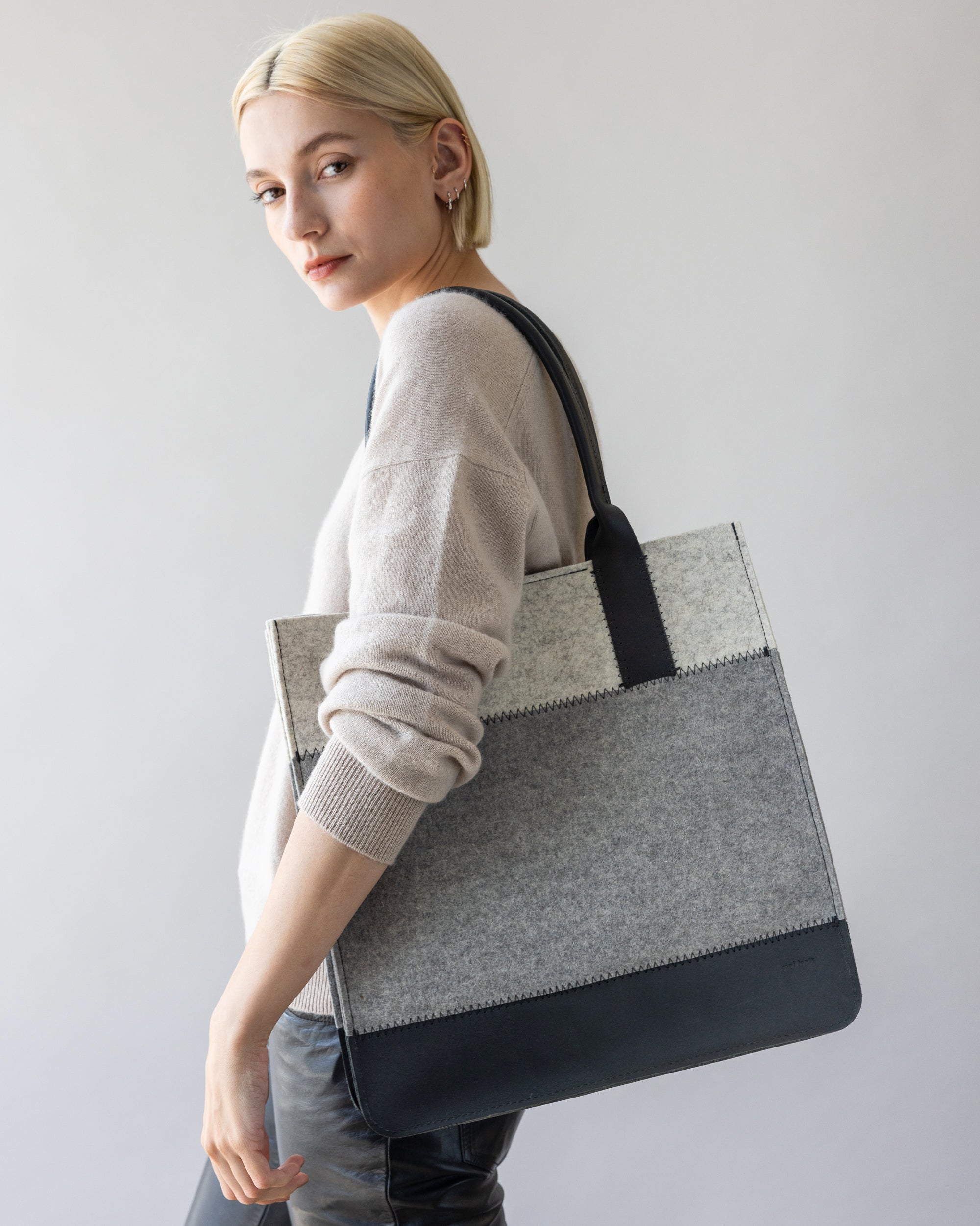 A person with short blonde hair poses with a durable graf lantz everyday bag made of Merino Wool on their shoulder, wearing a light sweater and dark pants against a neutral background.