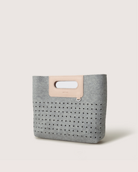 The graf lantz Asobi Bag is a minimalist clutch made from gray Merino wool, with a rectangular cut-out handle reinforced by a beige strap. The lower half features small, evenly spaced holes, all set against a light neutral background for an elegant and simple look.