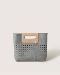 The graf lantz Asobi Bag is a minimalist clutch made from gray Merino wool, with a rectangular cut-out handle reinforced by a beige strap. The lower half features small, evenly spaced holes, all set against a light neutral background for an elegant and simple look.