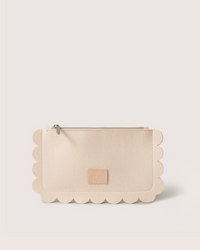 The Graf Lantz Deco Edge Pouch, crafted from beige Merino wool felt, features scalloped edges and a zipper closure. Its front is adorned with a small rectangular patch featuring embossed text for an elegant touch.