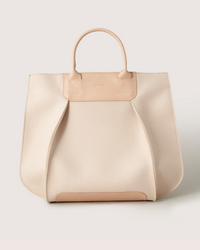 The Graf Lantz Frankie Tote features a cream-colored, minimalist design with a structured silhouette. Crafted from soft material, it includes sturdy light brown handles and subtle stitching details, perfectly blending style and functionality.