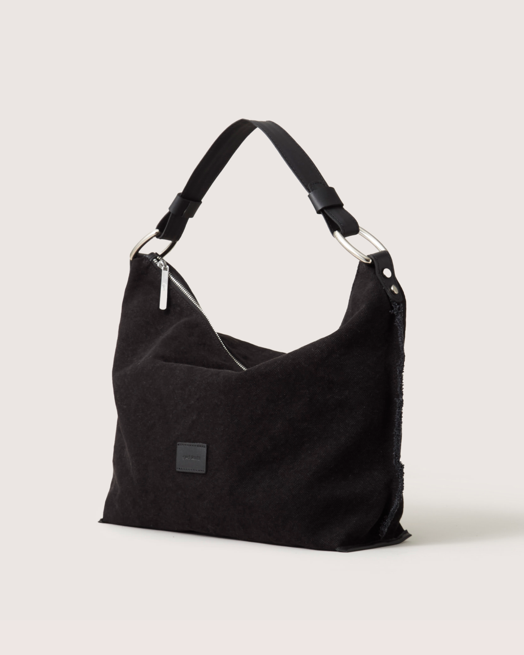 Part of Graf Lantzs Hana Bag Collection, this sleek black hobo features a wide leather shoulder strap, slouchy design, subtle textured fabric, silver hardware, and a front rectangular patch. Its neutral light gray background enhances its chic allure.