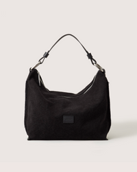 The Hana Hobo by Graf Lantz, a black suede shoulder bag with a single handle and metal accents, is shown against a plain white background. Part of the Hana Bag Collection, it features a subtle logo patch on the front.