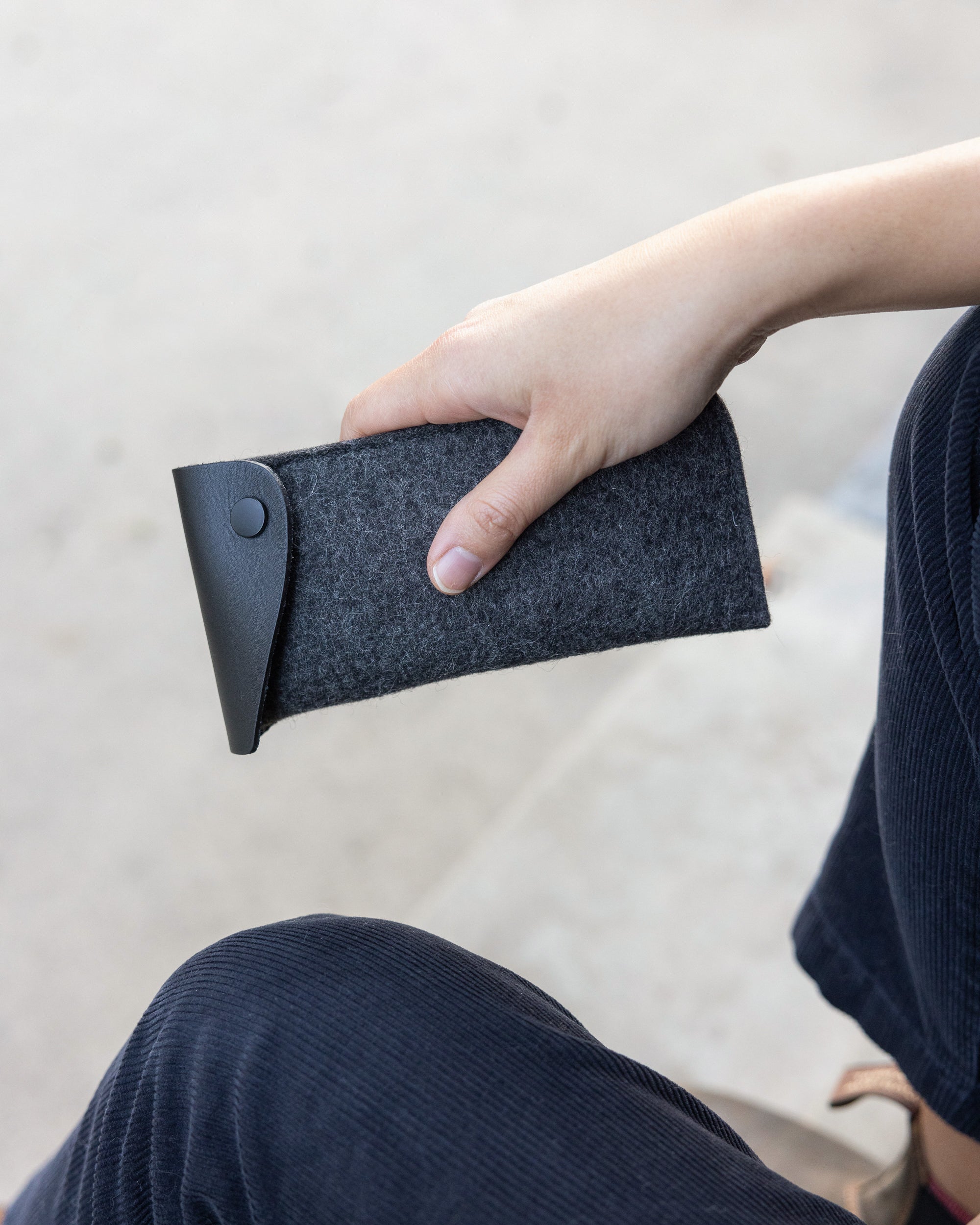 A person holds a chic graf lantz eyeglass sleeve made from gray Oeko-Tex® Certified felt with a black buttoned flap. They wear navy blue corduroy pants against a light gray concrete background.