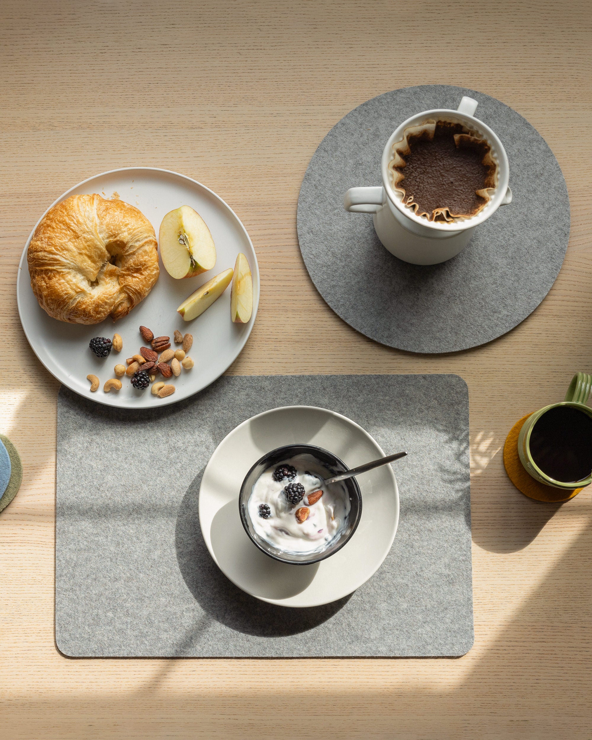 A breakfast scene includes a croissant, apple slices, nuts, and berries on a plate. A coffee mug rests on a graf lantz Merino Wool Placemat, known for its heat resistance. A yogurt bowl with berries sits on another placemat beside a small potted plant.