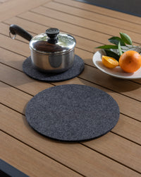 A metal saucepan with a lid sits on a heat-resistant gray felt trivet from graf lantz on a wooden table. Nearby is an empty gray felt trivet from graf lantz and a white bowl with an orange, lemon, and green leaves.