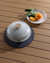 A ceramic dish with a heat-resistant wooden lid by graf lantz sits on a gray mat atop wood. Nearby, a white plate holds an orange, halved orange, and green leaves. Sunlight creates shadows across the setting.