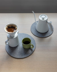 A minimalist coffee setup on a light wooden table showcases a white drip coffee maker and kettle resting on graf lantz heat-resistant Merino wool mats, with a green mug filled with freshly brewed coffee ready to be enjoyed.
