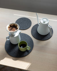 A tabletop features a graf lantz Merino wool trivet holding a white gooseneck kettle, coffee dripper with grounds, and white carafe. Nearby, a green cup sits on dark round mats as sunlight creates shadows across the light wood surface.