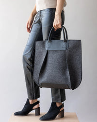 A person holds the graf lantz Frankie Tote, a large gray felt bag with black leather accents and handles. Dressed in black leather pants and heeled ankle boots, they display the totes structured elegance against a plain light background.