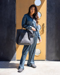 Dressed in a blue silk outfit, a person stands before a modern door with circular windows, holding a large gray Frankie Tote by graf lantz. Their slippers shuffle on the ground as sunlight casts shadows, highlighting their structured silhouette.