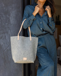 Dressed in a flowing, textured blue outfit, a person stands against a dark wall, effortlessly holding the leather handles of a large gray Hana Tote from graf lantz. The scene exudes casual elegance and style, highlighted by the refined details of the bag.