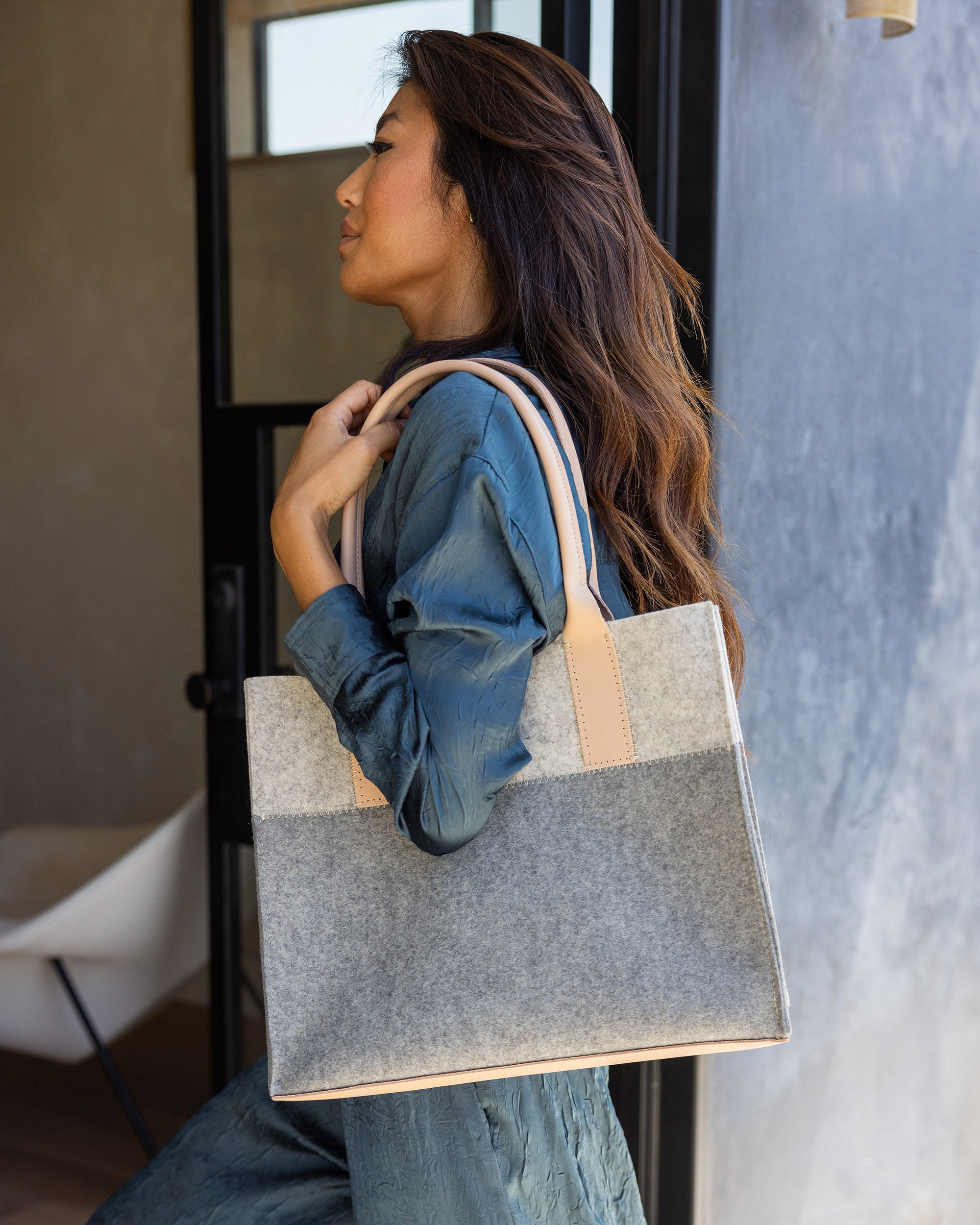 A person with long hair in a textured blue outfit stands indoors holding a stylish Jaunt Midi Tote by graf lantz, featuring tan handles and crafted from Merino Wool. They gaze upward slightly, with a modern chair visible in the background.