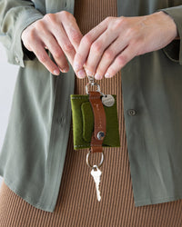 Standing woman holding a Merino Wool Pod Key Fob in Moss with brown leather accents in both hands in front of her, white background