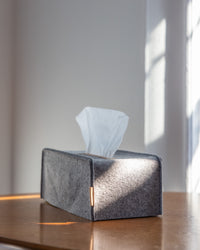 A graf lantz tissue box adorned in sustainable gray felt sits on a wooden surface, with a single white tissue pulled out while sunlight streams through a window, casting soft shadows behind.