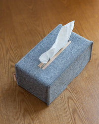 A gray felt tissue box cover by graf lantz, made from Merino wool with a beige leather strip at the opening, rests on a wooden surface. A partially pulled-out tissue enhances the texture contrast of this minimalist and sustainable design.