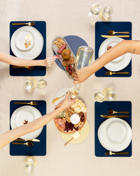 A top view showcases a dining table set for four with sustainable blue placemats and cozy merino wool accents from graf lantz. White plates, gold cutlery, and glasses of water complement the setting as two hands reach for a charcuterie board while a jug is poured during a toast.