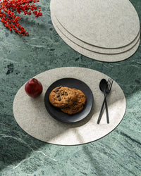 Cookies rest on a black plate beside a red apple, fork, and spoon on graf lantz Merino wool placemats atop a green marble table. Red berries accent the corner while sunlight casts shadows over this sustainable setup.