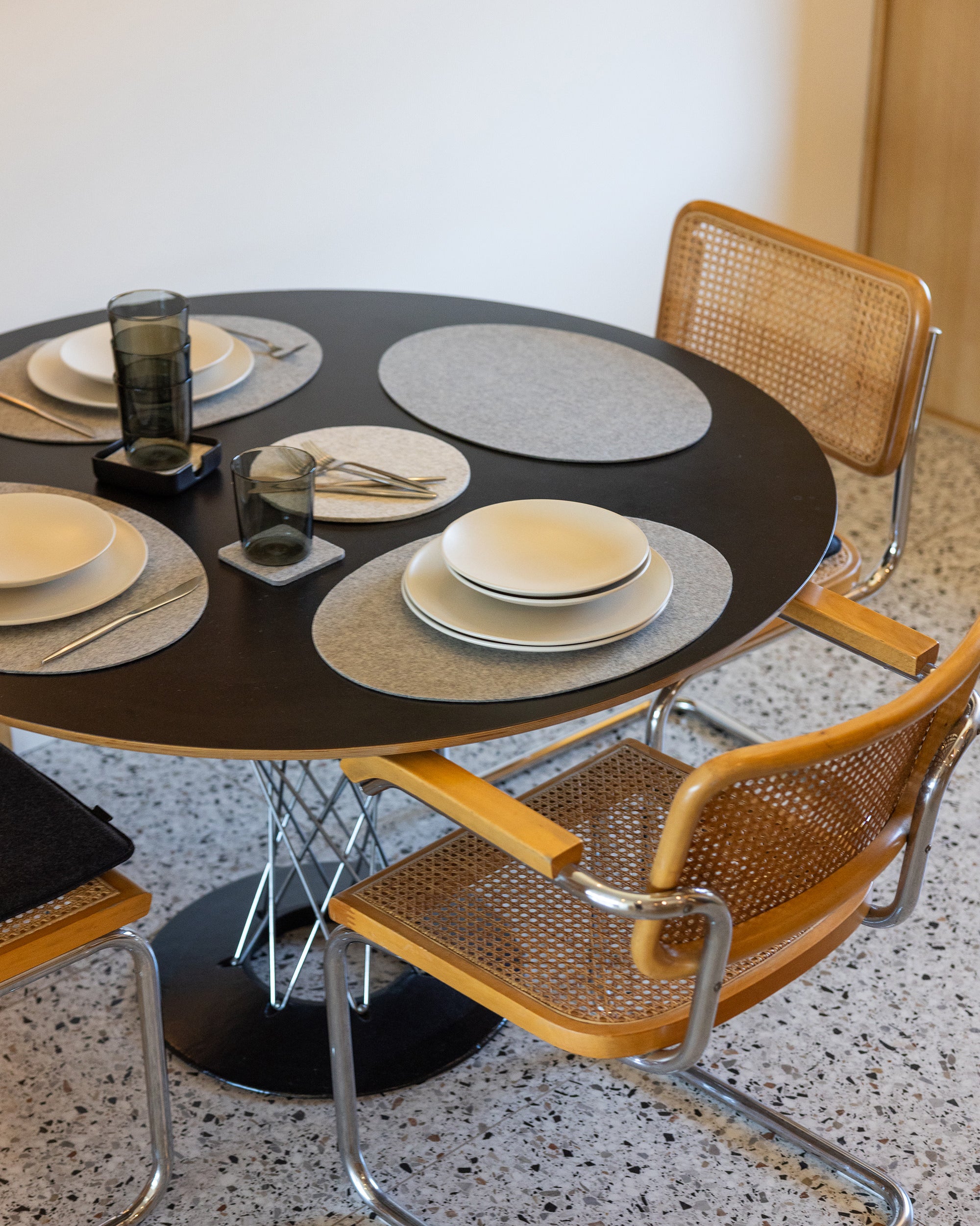 A modern dining setup showcases a round black table set with four place settings, each featuring a premium graf lantz Merino Wool Placemat. Complemented by white plates, black rectangular napkins, dark glasses, and silverware, the ensemble is completed with chic wicker and metal chairs.