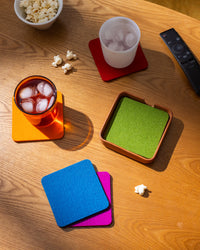 A wooden table is adorned with graf lantz colorful German Merino wool coasters, holding two iced drinks on orange and red coasters. A bowl of popcorn and scattered kernels sit beside a TV remote on a green coaster, while bright lighting artfully casts shadows, enhancing the sustainable décor.