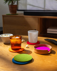 A wooden table holds a glass of iced drink and a hot beverage in a white mug, both on graf lantz Merino wool coasters. A Kobon leather tray from graf lantz cradles popcorn, while the TV remote and extra coasters are scattered around as sunlight casts gentle shadows.