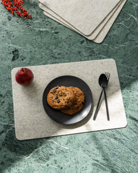 A black plate with three chocolate chip cookies sits on a rectangular, graf lantz designer-inspired placemat. Nearby, a red apple and utensils are positioned beside the plate, with bright lighting casting shadows that highlight the green marble surface below.