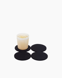 A small white cup filled with ice rests on a sleek black Bierfilzl Merino Wool Round Coaster. Three more identical coasters, soft as beer felt, are artfully arranged nearby on a pristine white background.