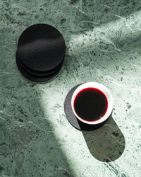 A white cup with dark red liquid rests on a Bierfilzl Merino Wool Round Coaster on a green marbled surface. Beside it is a stack of three black coasters. A sunlight streak casts contrasting shadows.
