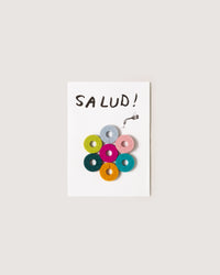 A postcard from graf lantz features SALUD! at the top with a small faucet illustration, and a pyramid of seven colorful Merino wool yarn donuts, ideal as a distinctive host gift.