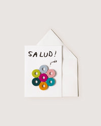 The graf lantz greeting card features an arm holding a drink and colorful donut shapes in a floral design with SALUD! exclaiming joy, making it perfect as a host gift. It comes partially visible from a white envelope against a light backdrop.