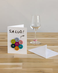 A graf lantz greeting card with colorful donuts and Salud! sits on a wooden table. Nearby, a white envelope and an empty wine glass with a pink marker add charm, all against a plain light-colored wall—ideal as a thoughtful host gift.