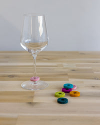 A wine glass with a pink felt ring as a marker around its stem sits on a wooden table. Nearby, graf lantzs colorful felt rings in blue, green, and orange—crafted from soft Merino wool—are scattered, making them the perfect playful host gift.