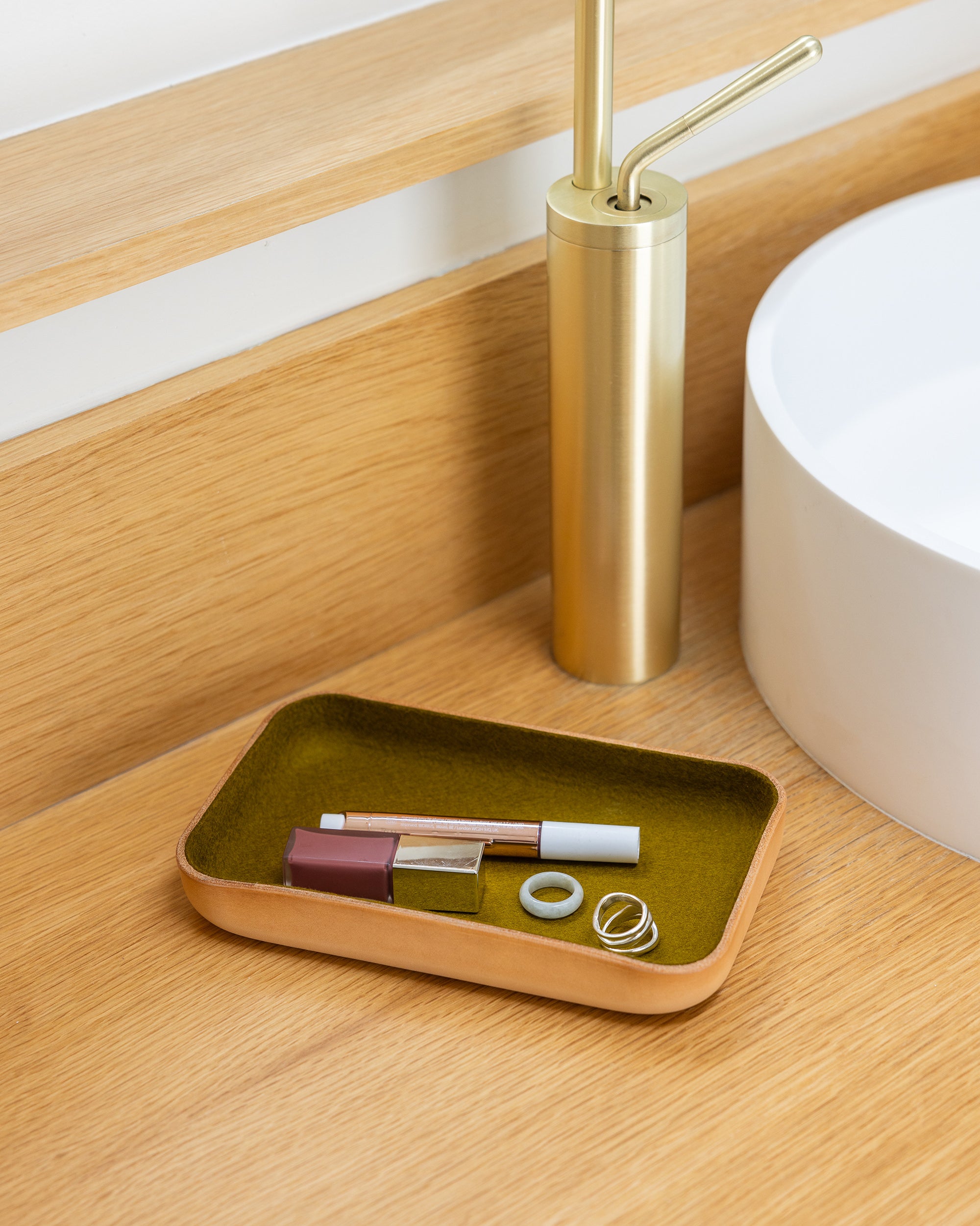 On a light wooden surface, a graf lantz wooden catchall tray neatly holds makeup items and two rings. Nearby, a gleaming gold pump dispenser sits beside part of a white sink, adding an elegant touch to the serene setting.