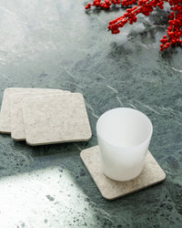 A white frosted glass rests on a square, light gray graf lantz merino wool coaster on a dark marble surface, with three more coasters stacked nearby. Red berry sprigs in the upper right corner add a festive touch to the home décor.