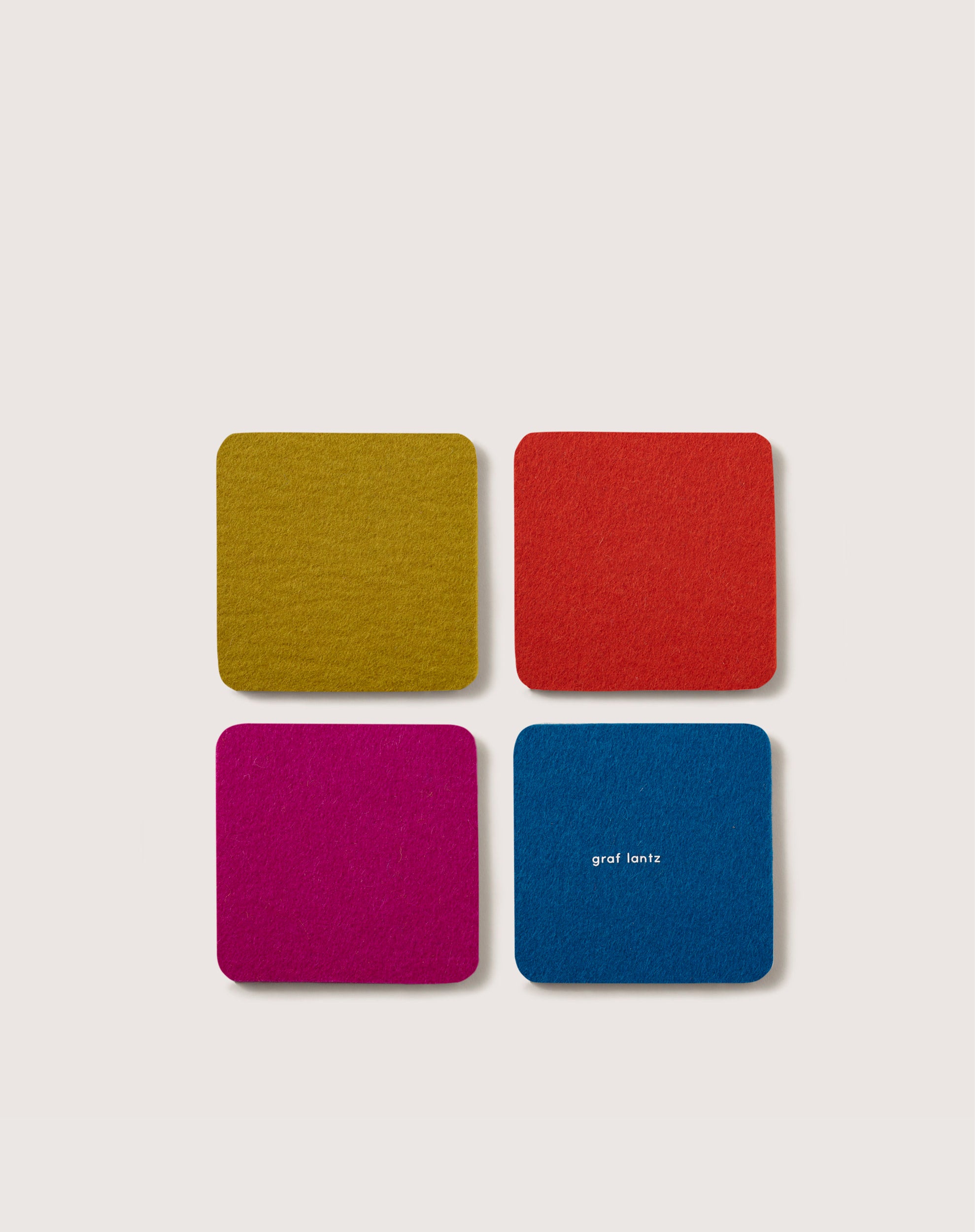 These four square felt coasters, crafted from premium Merino wool in vibrant hues of yellow, red, magenta, and blue (with graf lantz in white on the blue coaster), add a stylish touch to home décor or serve as essential beer mats. They are displayed neatly on a light backdrop.