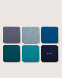 A set of six square felt coasters made from premium Merino wool in blue and teal hues are displayed in two rows on a light background, with one coaster featuring the brand name graf lantz, showcasing their meticulous handcrafted detail.