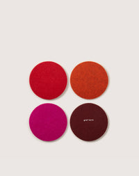 A square grid of four round graf lantz coasters in red, orange, pink, and burgundy German Merino wool sit on a light background. One coaster displays the brand name, merging style with sustainable home décor.