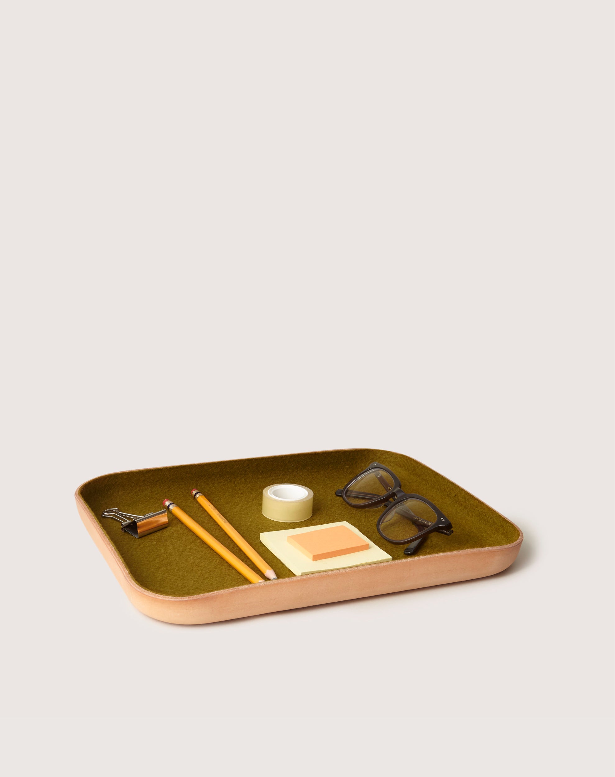 A graf lantz wooden catchall tray with an olive green interior holds glasses, yellow pencils, a tape roll, a binder clip, and sticky notes. Its minimalist design contrasts beautifully against the plain white background.