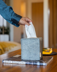 A hand pulls a tissue from a graf lantz gray Merino Wool cube tissue box on a wooden table. The Oeko-Tex® Certified cover is set on a book titled MARK BRADFORD, with a softly lit room blurred in the background.