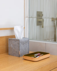A modern bathroom with a wooden countertop features a graf lantz Merino Wool tissue box cover beside a wooden tray holding lipstick and mascara. This elegant scene is set against the backdrop of a mirror and shower, merging natural elegance with contemporary flair.