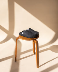 A pair of dark gray slipper shoes by graf lantz, made from sustainable Merino wool felt, rests on a wooden stool with a beige wall and cross-shaped shadows in the background. Warm lighting enhances the cozy ambiance.