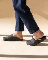 A person in dark blue pants walks, displaying gray Merino wool felt slippers from graf lantz. The plain cream background with sunlight casting soft shadows enhances the allure of sustainable footwear.