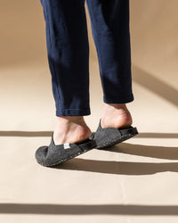 Someone in dark blue pants and graf lantz gray slipper shoes, made from Merino wool felt, stands on a beige surface. The slippers are slightly elevated as sunlight elegantly casts shadows on the floor.