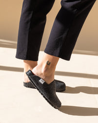 A person is depicted wearing graf lantz black sustainable slip-on shoes and black pants, with a small horseshoe tattoo on their ankle. Sunlight casts shadows on the beige floor, enhancing the elegant simplicity of their footwear choice.